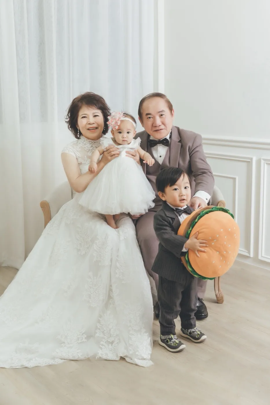 Grandparents in a white lace gown and a grey suit seated in the studio with their baby granddaughter and toddler grandson holding a plush burger