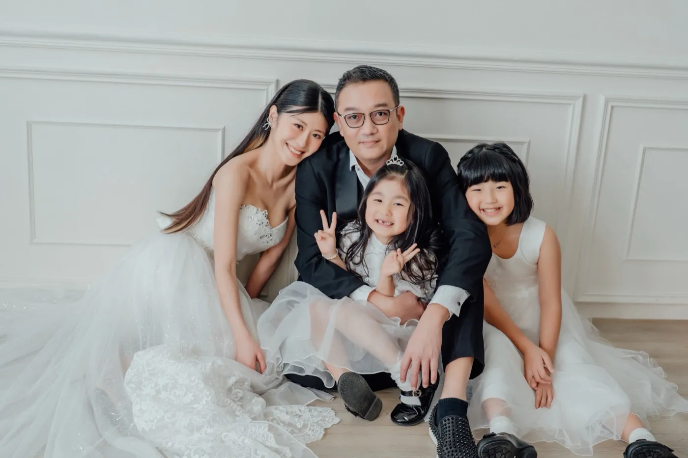 A family in white against a white panelled wall: mum in a strapless gown, dad in a black suit and their two daughters in white dresses