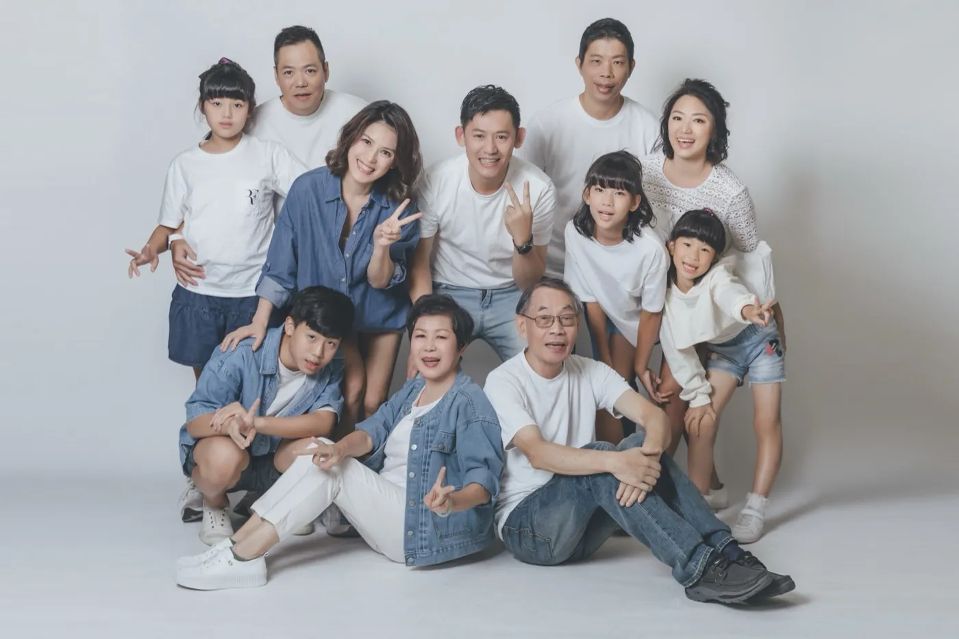A large three-generation family in white T-shirts and denim, grandparents seated in front, everyone smiling and making peace signs