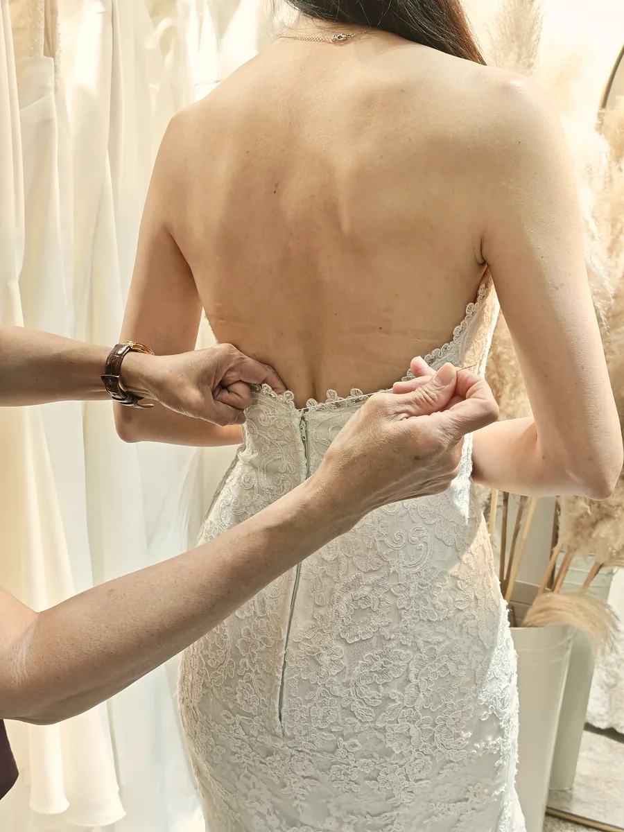 A bride’s back as her consultant fastens a lace gown